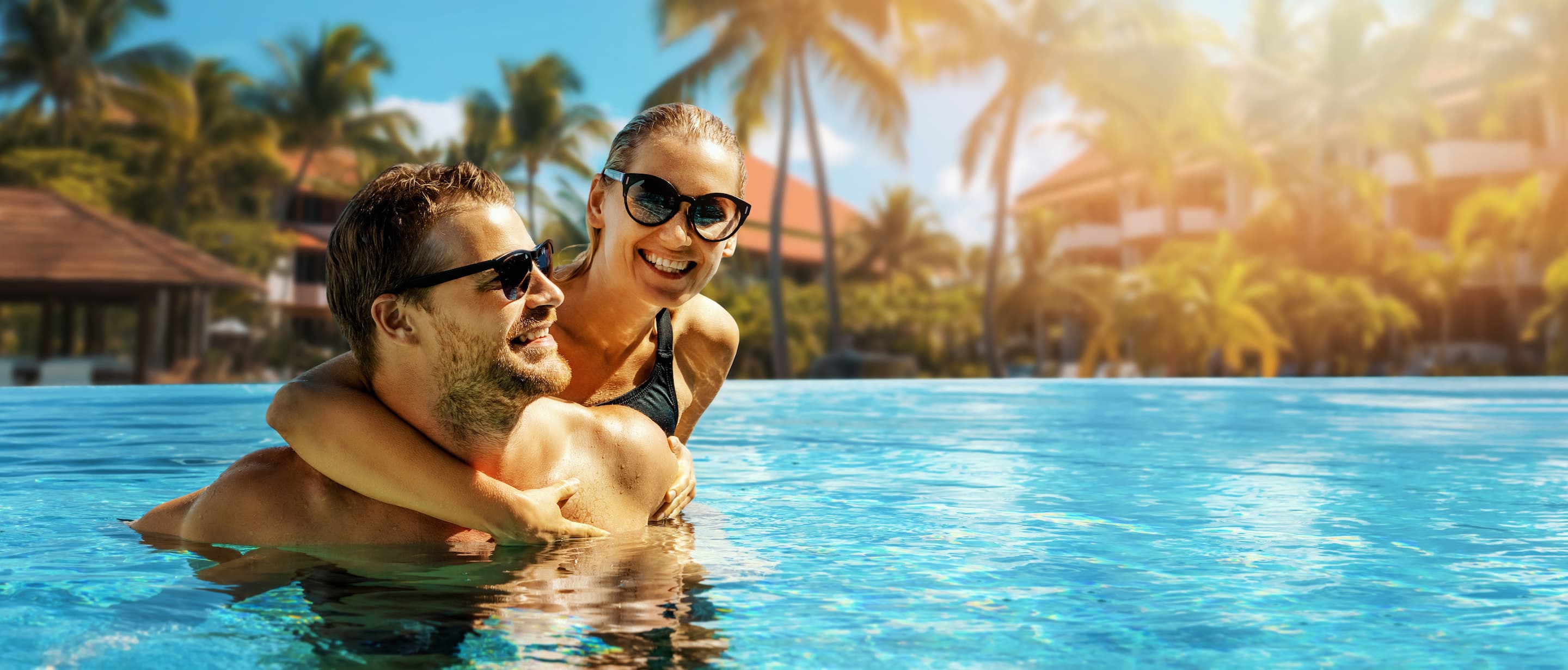 A smiling couple wearing sunglasses enjoys a swim in a pool, surrounded by palm trees and resort buildings under bright, sunny weather.