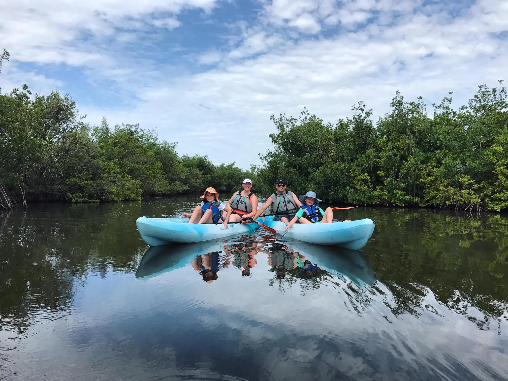 Four people sit in two blue kayaks on calm water surrounded by green trees and under a partly cloudy sky.