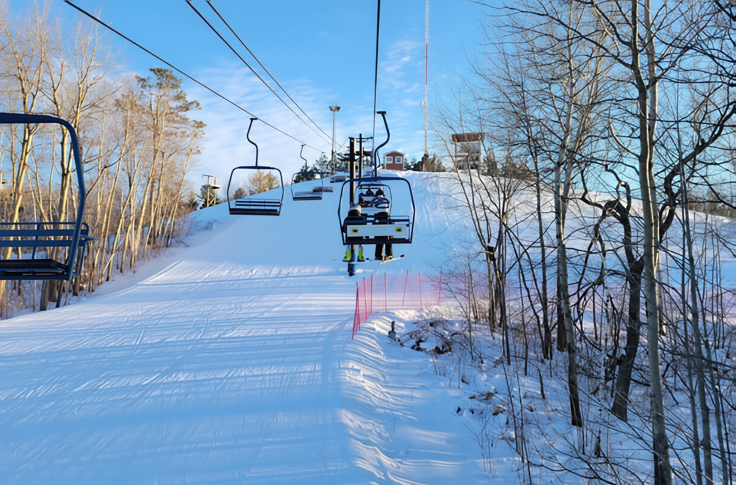 A ski lift carries people up a snowy slope lined with bare trees on a clear, sunny winter day.